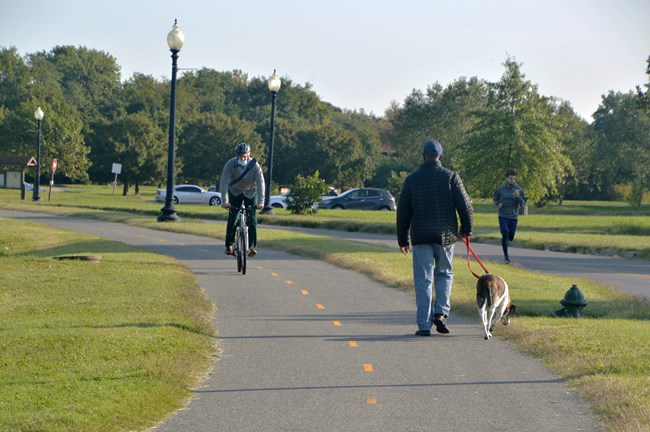 A pedestrian walks their dog along the Anacostia Riverwalk trail, as a cyclist and jogger pass by.