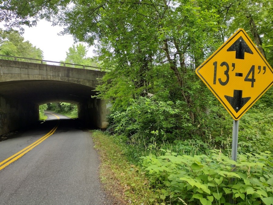 A road on the left of the image, with a bridge about 100 feet in the distance. On the right side, a 13 feet 4 inch clearance sign in the foreground with overgrown vegetation in the background along the roadway.