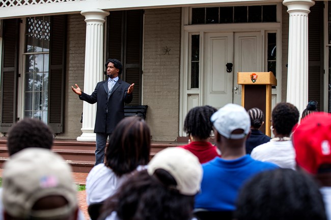 A historical interpreter portraying Frederick Douglass stands on the front porch of a large historic house, speaking with expressive gestures to an audience seated on the lawn.