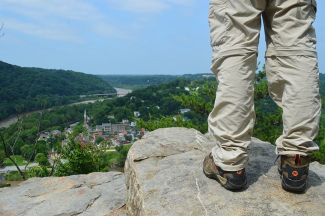Hikers legs standing on a rock on the Maryland Heights Trail. The town of Harpers Ferry is visible below.