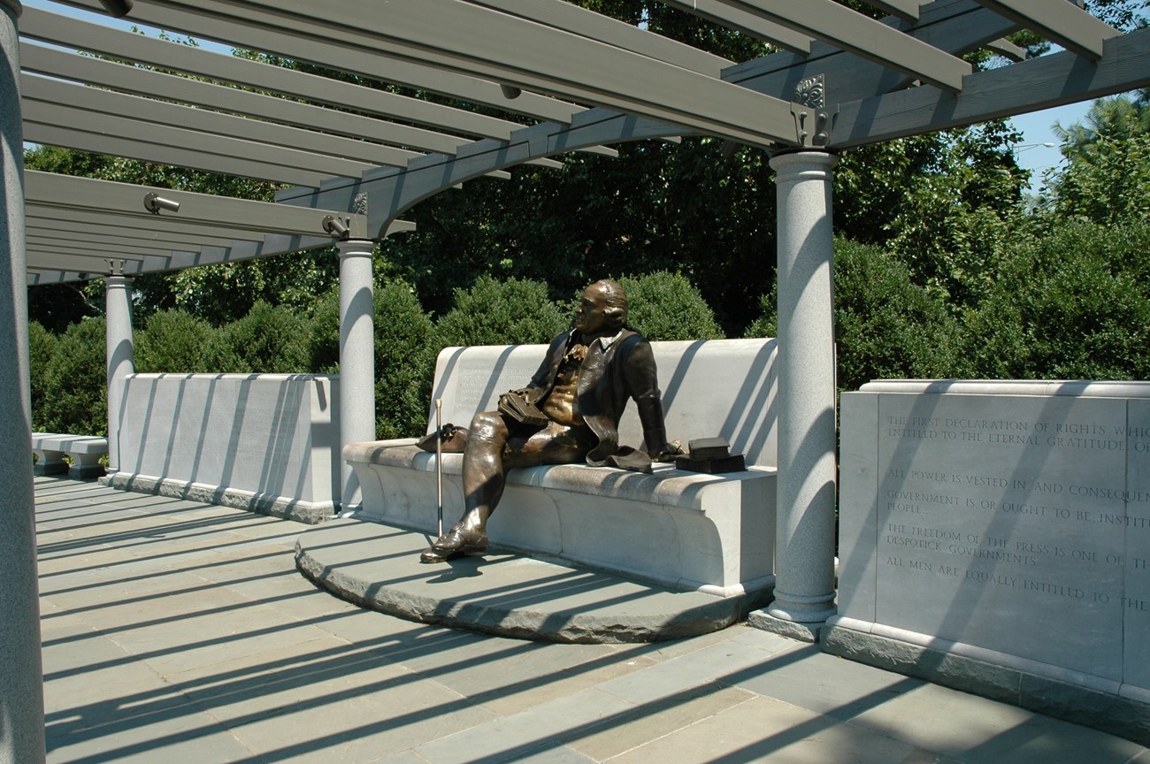 A bronze statue of George Mason seated with his legs crossed on a stone bench.