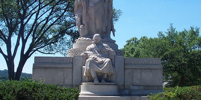 Statue of man sitting down on a pedestal with statue above him surrounded by bushes and trees.