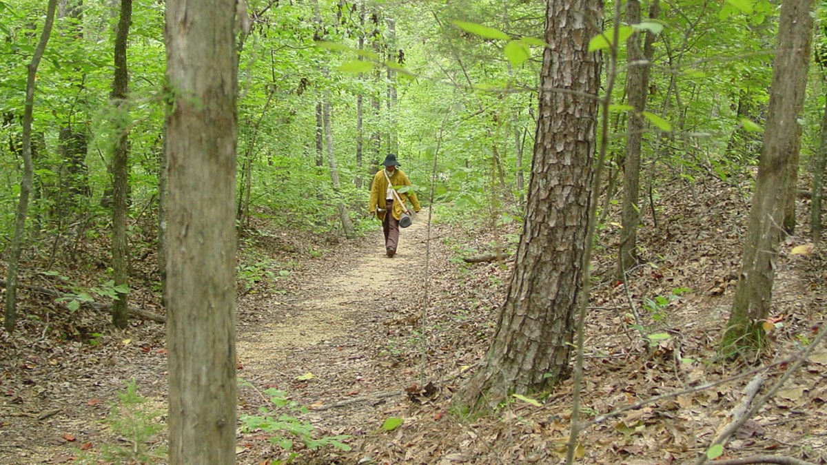 A man in pioneer clothing walking on a trail through the woods.