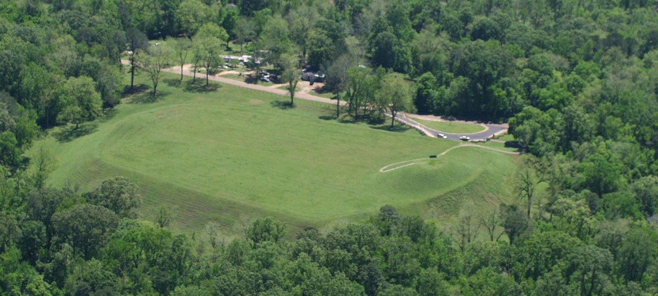 Aerial view of Emerald Mound. Mound is covered in mowed green grass and a forest filled with green leaves surrounds the mound site