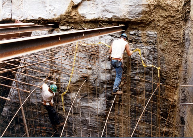 Two men in hard hats and jeans descend into a stone hole down a metal rebar grid