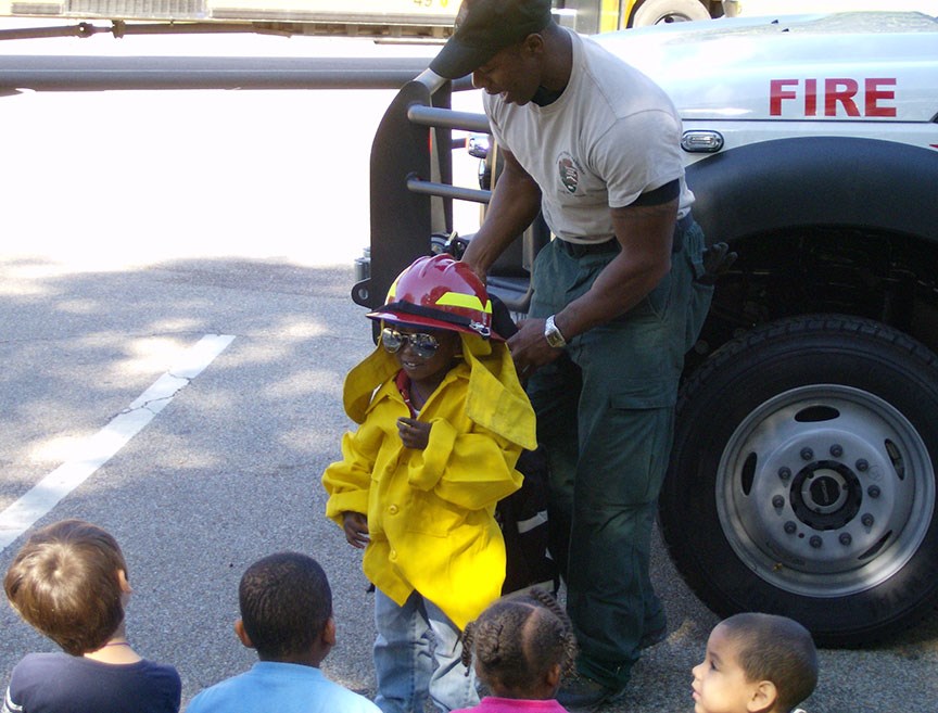 A firefighter helps a young child dress in firefighter gear.