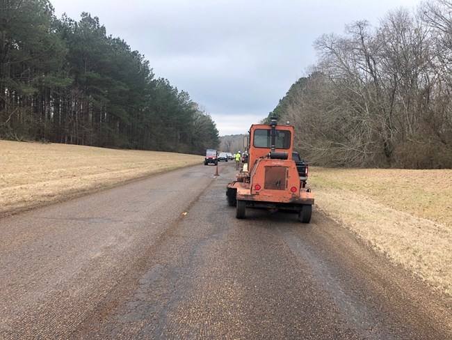 An orange pavement groover occupies one lane while a lead vehicle leads oncoming traffic toward it.