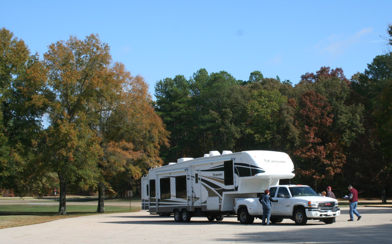 Campers head out after a stop at Parkway Visitor Center in Tupelo, MS (MM 266) A large white pickup truck towing a large 5th wheel travel trailer is parked in a parking lot. Four people walk toward the vehicle, one looking at a brochure. The lot is surrounded by trees with fall foliage.