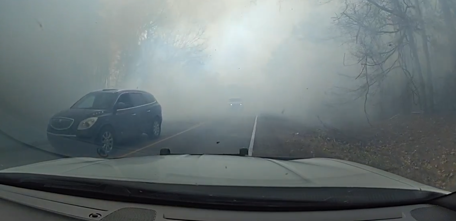 A law enforcement dash cam view of a smoke filled road with a black car passing northbound and a white truck in the distance.
