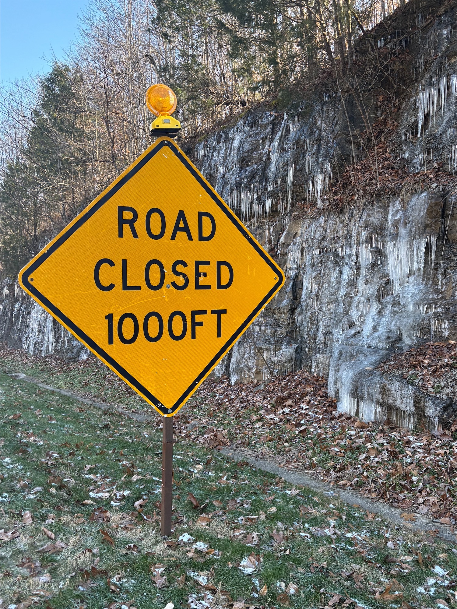 A yellow warning sign with blinking light at the top states "Road Closed 1000FT". The sign stands in a grassy area with a rocky wall and bare trees behind it.