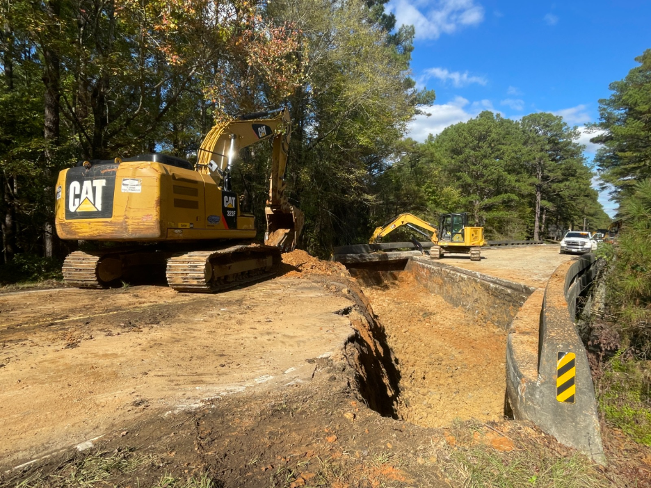 Two excavators on opposite sides of a gap in the bridge. The road is entirely dirt and clay.