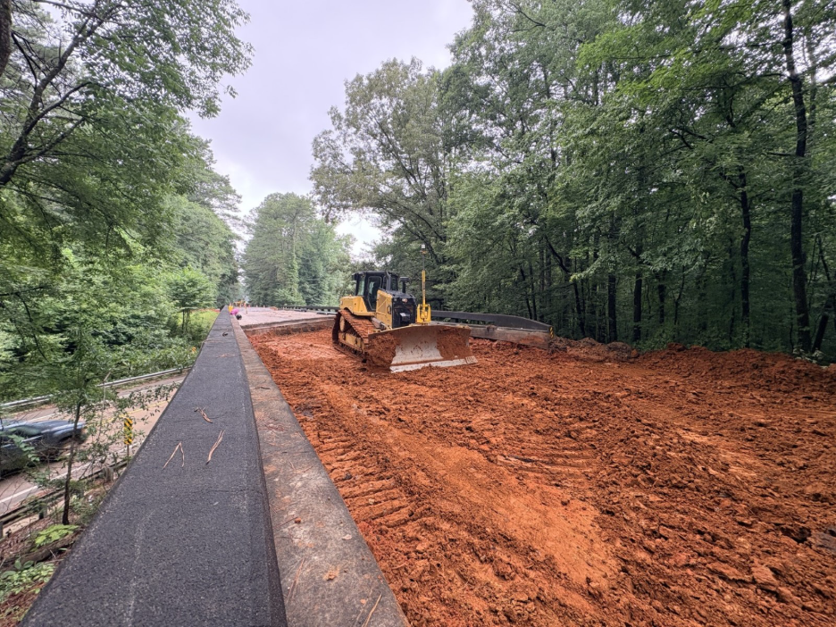 A yellow bulldozer sits on a wide, freshly excavated dirt section of the Natchez Trace Parkway, surrounded by tall, leafy green trees. The construction area is bordered by a paved road edge, indicating active roadwork.