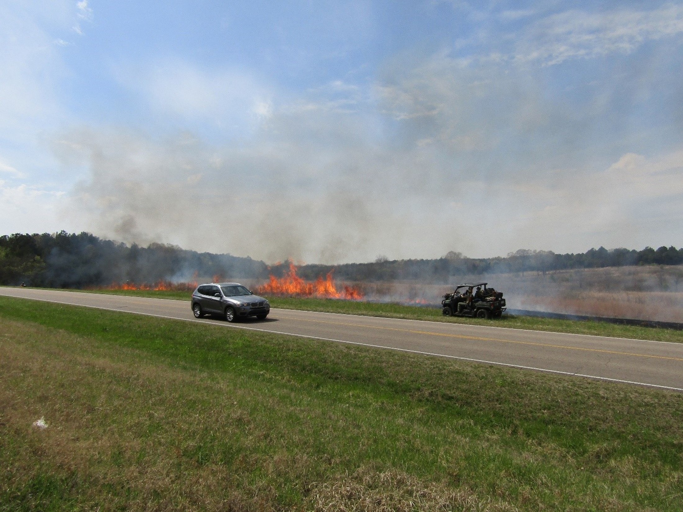 MicrosoftTeams-image (15) Fire on the side of the road with a vehicle driving by. Smoke is in the air.