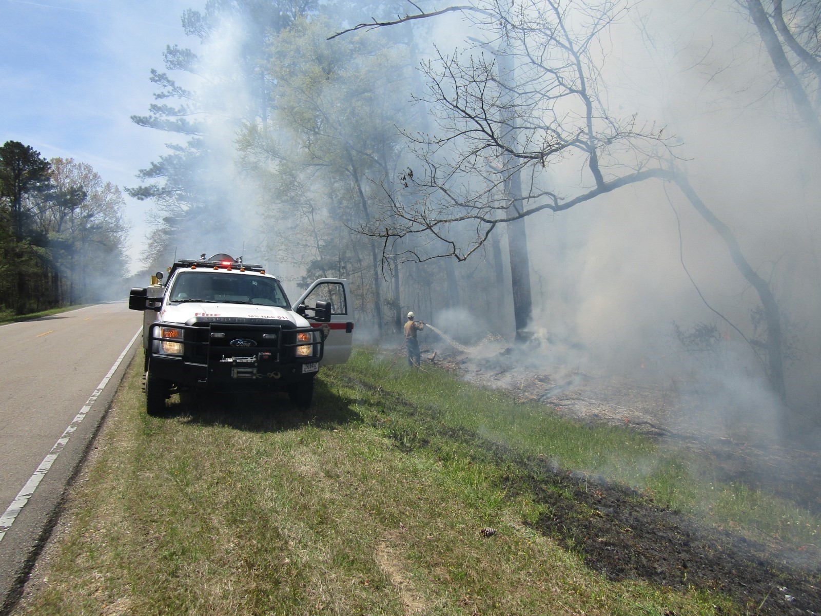 A single firefighter sprays water on a fire at the line where the forest meets the grass. Smoke fills the woods. A fire engine is parked on the grass and with the driver side door hope. A single lane of the parkway is visible in the left of the frame. A single firefighter sprays water on a fire at the line where the forest meets the grass. Smoke fills the woods. A fire engine is parked on the grass and with the driver side door hope. A single lane of the parkway is visible in the left of the frame.