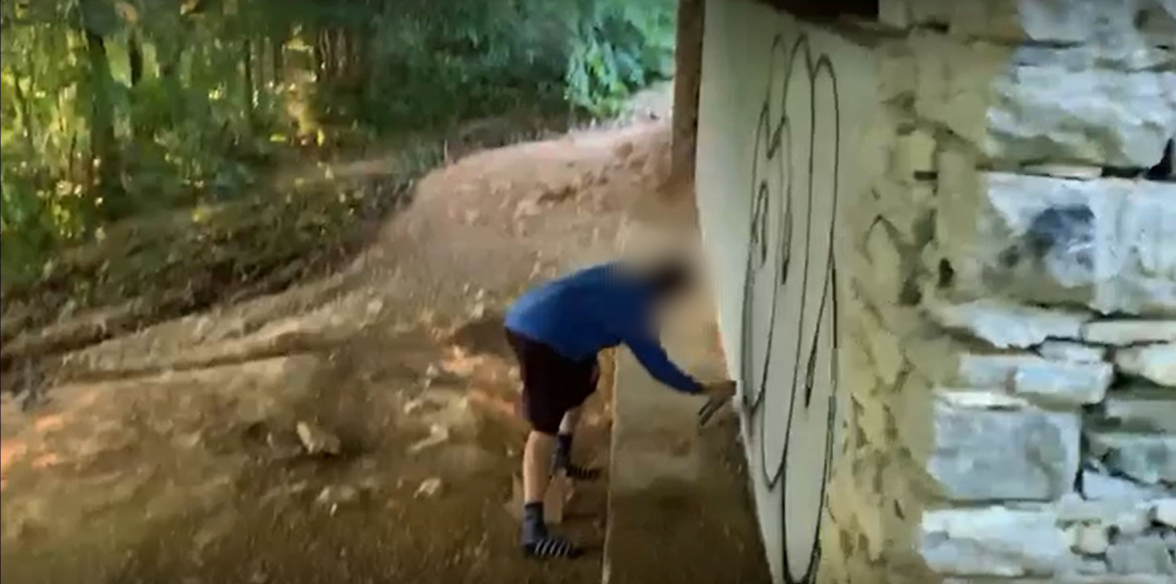 Man in long sleeve blue shirt and dark shorts standing on dirt spray paints a white wall with black lettering.