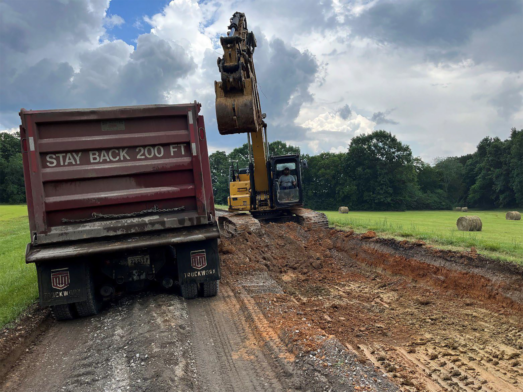Removal of failing materials from existing roadway A large dump truck and backhoe sit side by side on what was once a two lane road that is now dug down to the dirt level below the road. Deep tire and tractor tracks cover the dirt road bed. The outline of the backhoe operator in a grey shirt is visible in