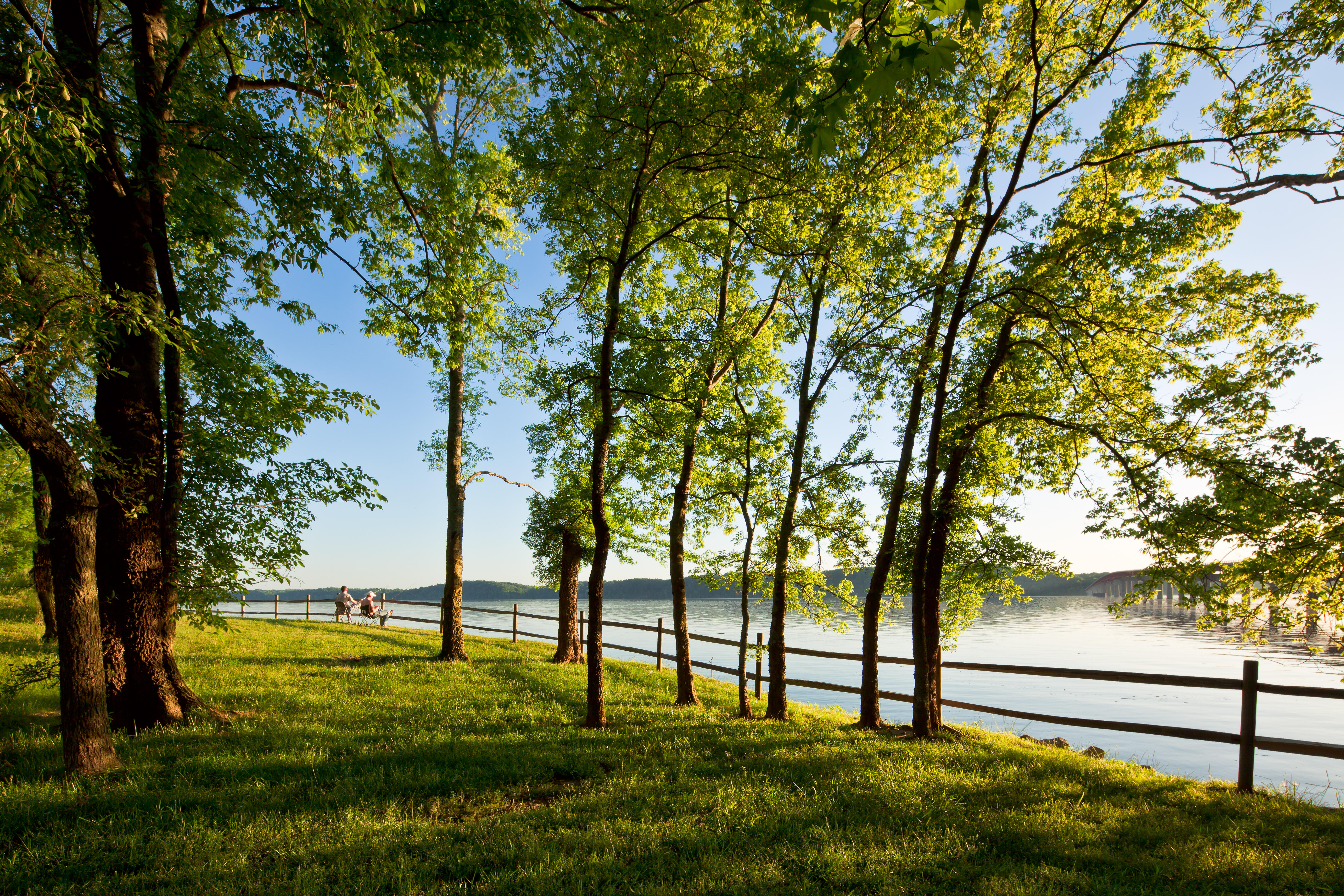 Water Route Overlook, Milepost 328.7 Green tree covered grassy area with wooden railing lining a river. Two people sit near the railing in the distance.