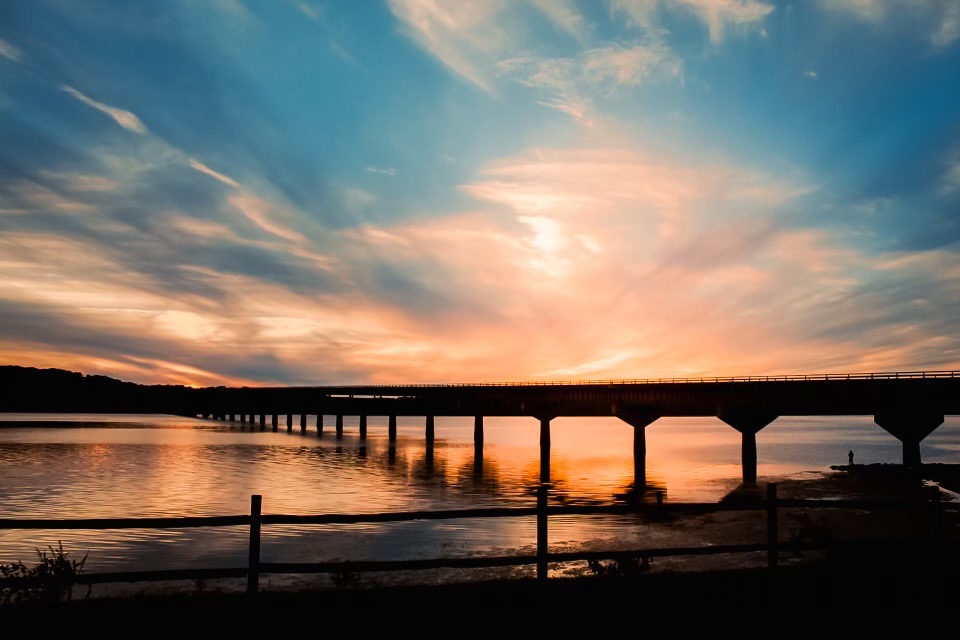 The John Coffee Memorial Bridge replacement EA is now open for public comment. The silhouette of a bridge at sunset spanning a wide river. The bridge stands on singular pilar supports. The sky is pink and blue with swirling clouds.