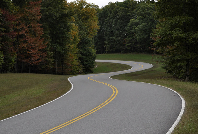 Natchez Trace Parkway encourages motorcycle riders to travel safely: “Different Road, Different Ride” An empty two lane road curves in the shape of the number three. The road is lined with short green grass and tall green trees that are just beginning to show signs of fall with scattered yellow and copper foliage on the left side of the road.