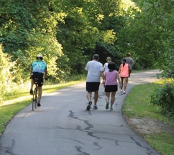 Chisha Foka Multi-Use trail A group of five people walk for exercise on a paved trail. A bicyclist passes them on the left of the trail. The trail is lined with green grass and trees.