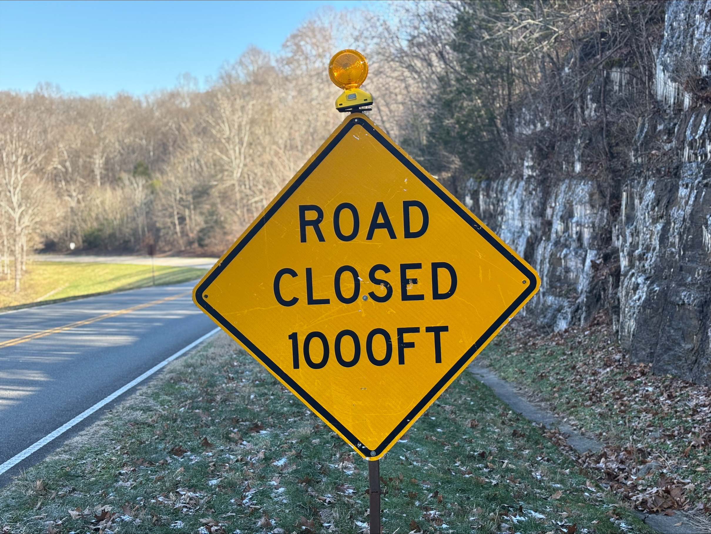 Yellow road closure sign stands in front of winding road through trees.