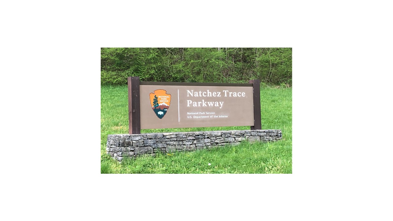 A brown rectangle sign with a National Park Service Arrowhead and the words Natchez Trace Parkway National Park Service U.S. Department of the Interior in white lettering. The sign is on a grey flat stone base with bright green grass all around it.