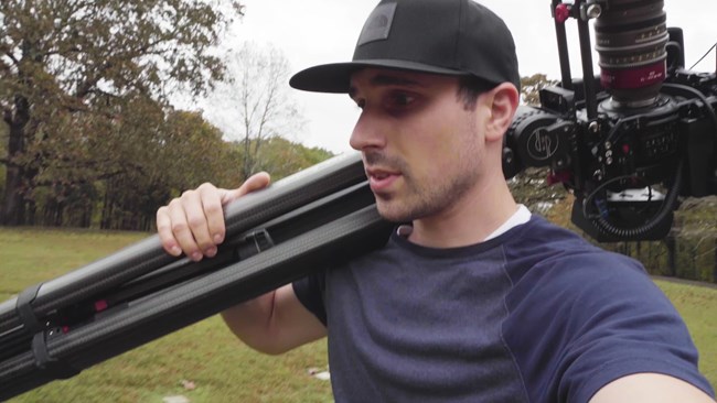 Man carrying a large tripod with a camera on his shoulder.