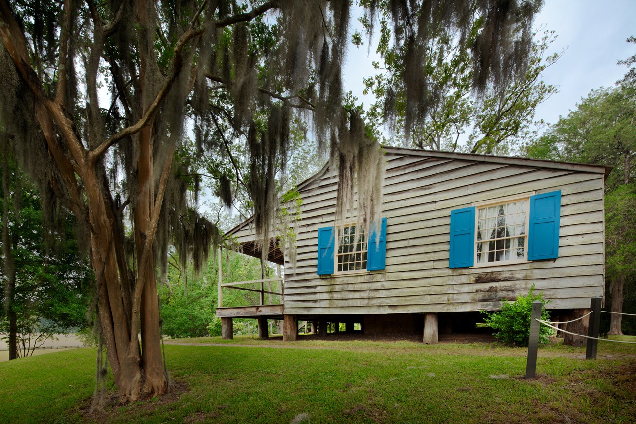 Side view of Mount Locust Inn and Plantation. Wooden siding on teh house is gray in color with light blue shutters on the two windows facing the viewer. The front porch is to the left and is covered and a wood railing goes around the entire porch.
