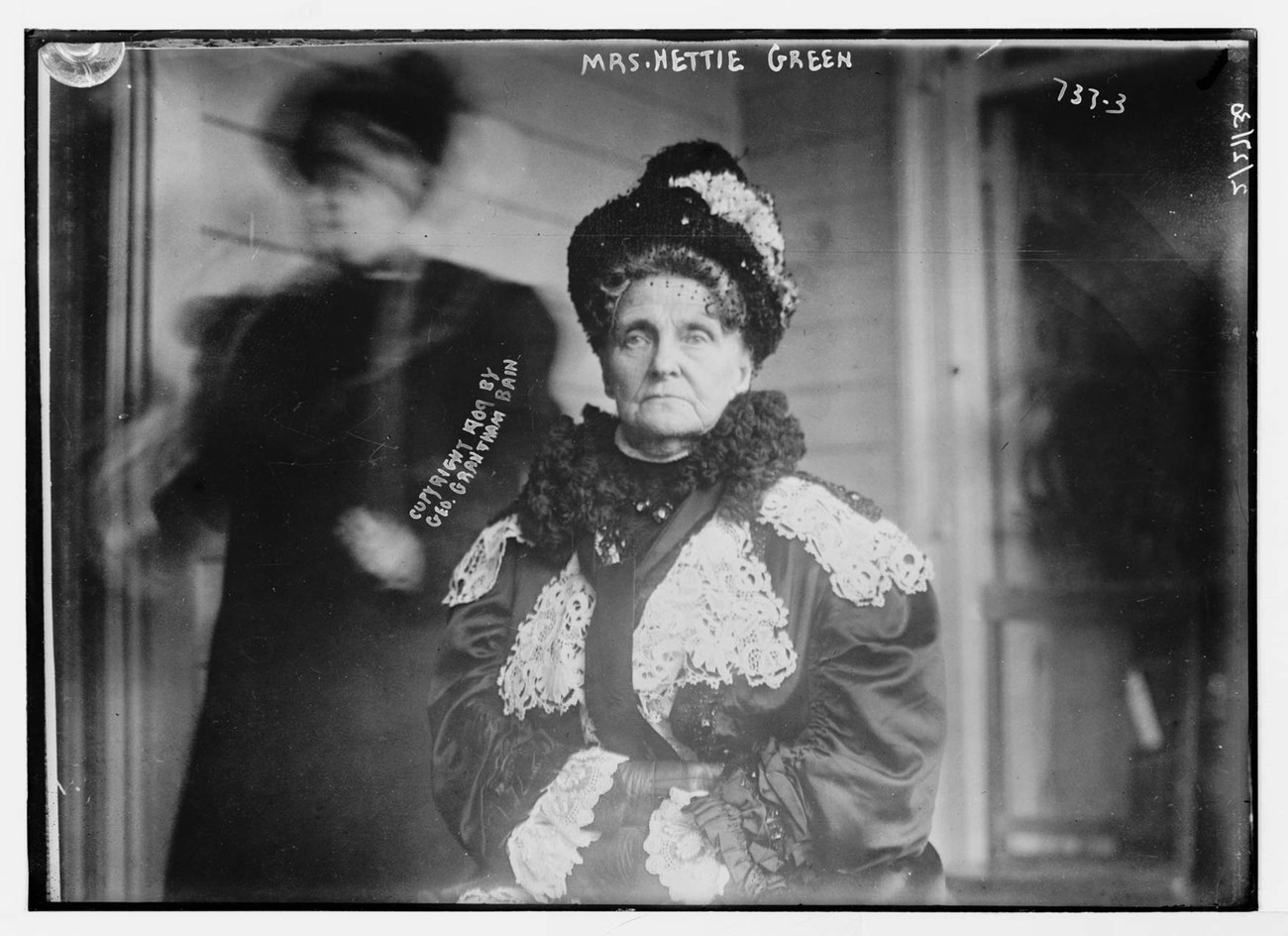 Black and white photo of Henrietta Green. Green sits with her arms folded, wearing a doily covered coat and hat.