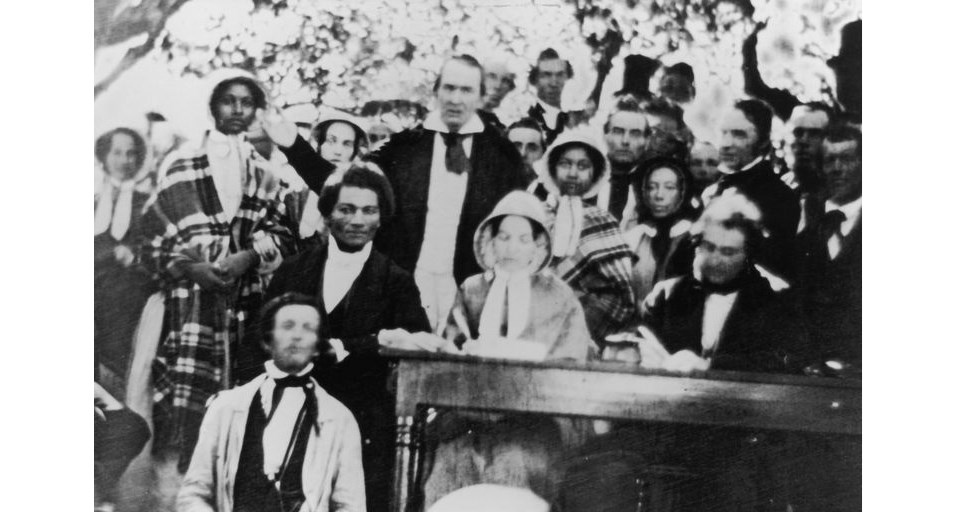 Black and white photo of Frederick Douglass leaning against a table among a crowd during an anti-slavery convention.