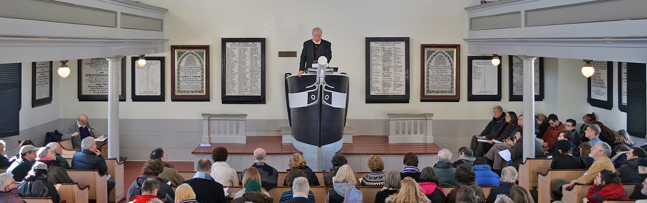 A full crowd listens as a religious leader stands at the prow-shaped pulpit.