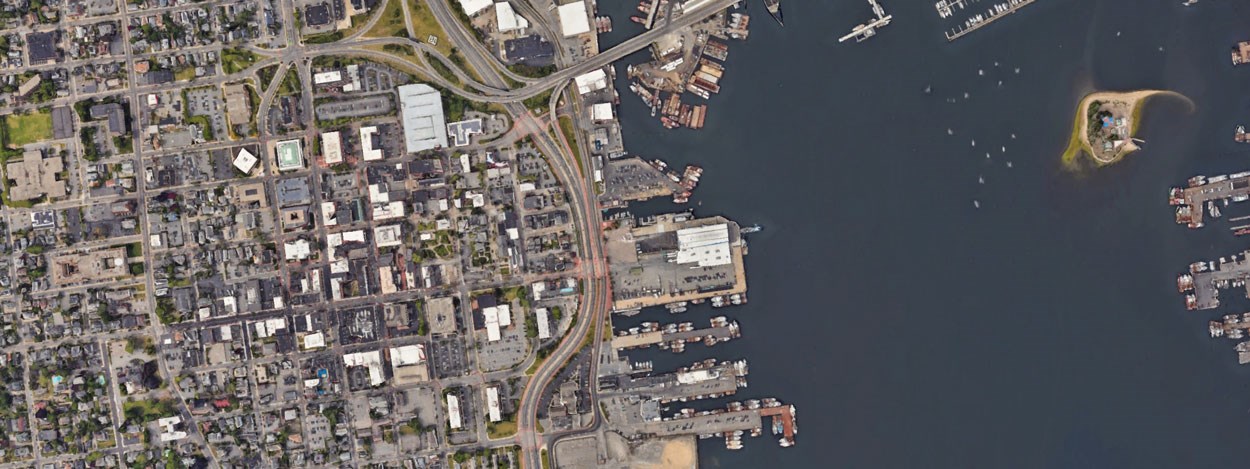 Aerial view of downtown New Bedford, Massachusetts, showing the intersection of highways and roads near the waterfront. The image highlights docks, piers, and an island in the harbor, with surrounding buildings and streets laid out in a grid pattern.