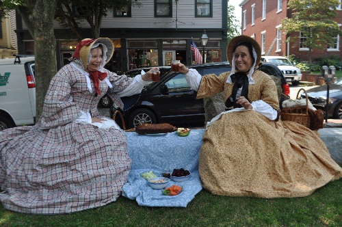 The 1850s ladies, in period dress are seated at a picnic spread with fruits, vegetables and breads.