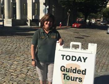 woman in volunteer shirt stands outside by a sign offered guided tours