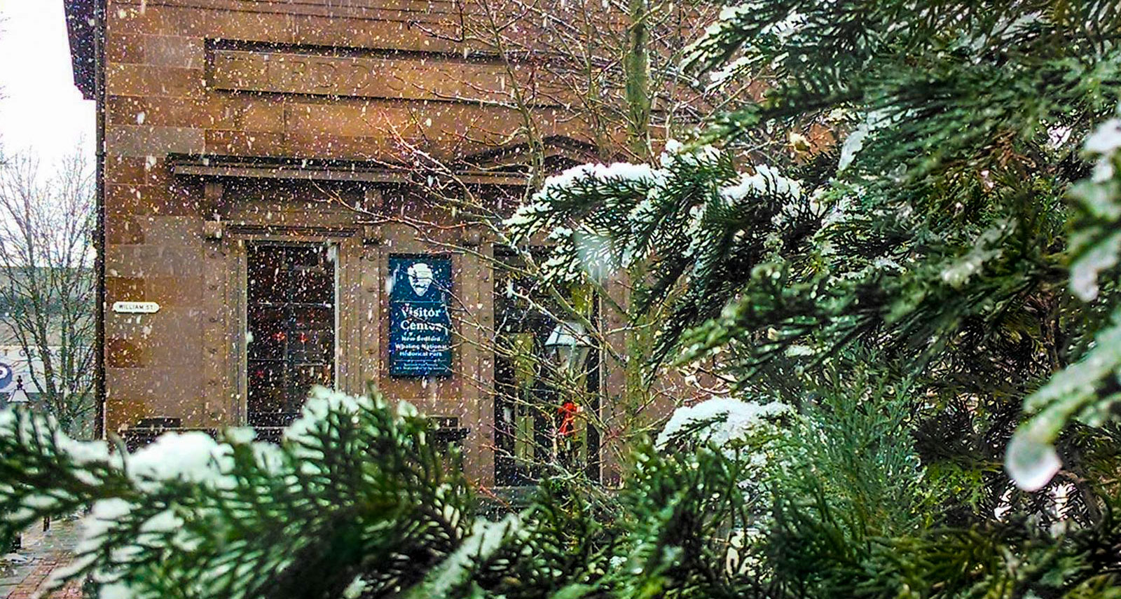 Snow falls on the New Bedford Whaling National Historical Park Visitor Center, framed by snowy evergreen branches.