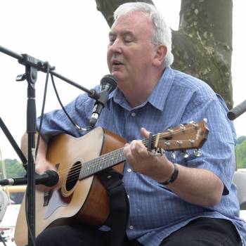 Robbie O'Connell an older man plays a guitar and sings into a microphone