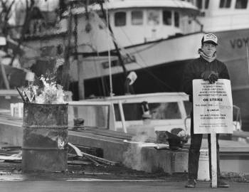 young man holding an "on strike" placard stands next to a fishing boat at a dock