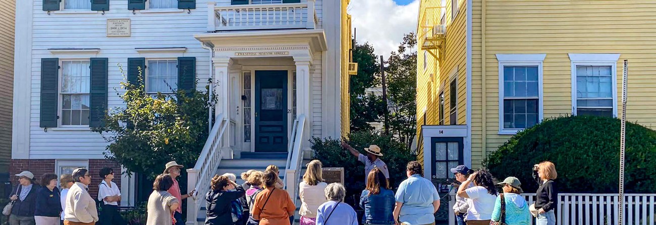 A park ranger leads a group of visitors on a guided walking tour outside the white, historic Nathan and Polly Johnson House in downtown New Bedford. The ranger gestures toward a plaque while the group listens attentively.