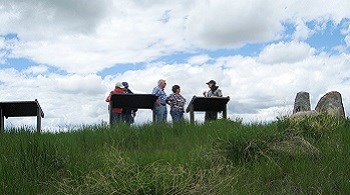 A park ranger talks to four visitors in front of wayside signs on a summer day.