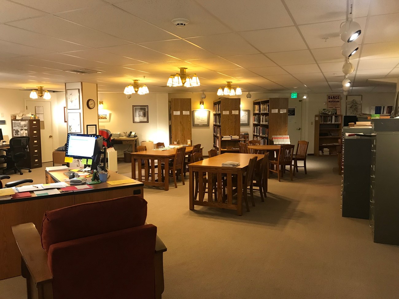 A room with many tables and chairs is empty. There are many file cabinets and bookshelves with books.