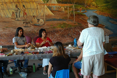 Cultural Demonstrators Two native women sit behind a table covered with materials for weaving. Three women sit on the opposite side of the table watching and listening to the demonstrators.