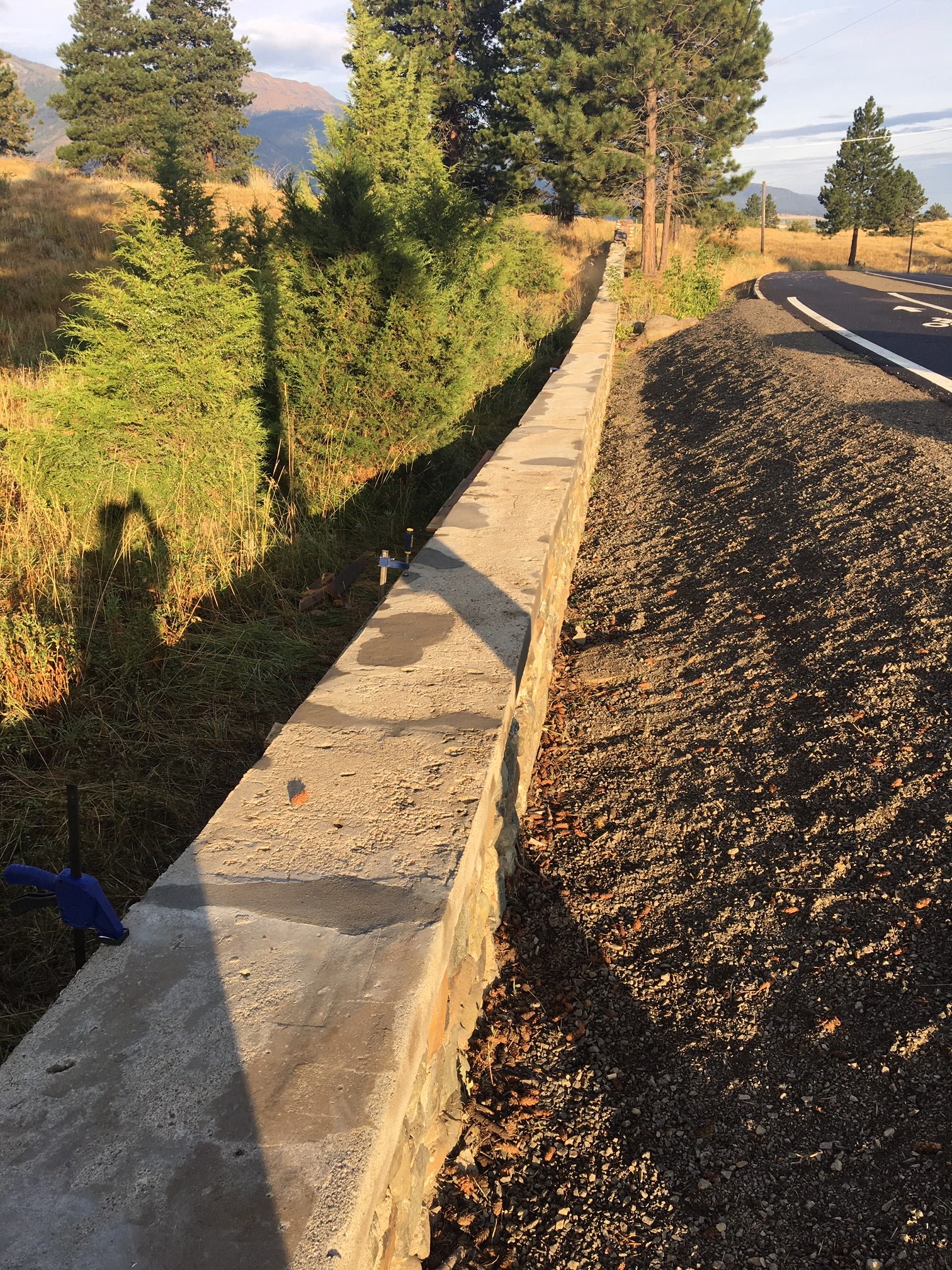 A grey concret-topped wall stands next to a road and open land in the sunset sunlight.