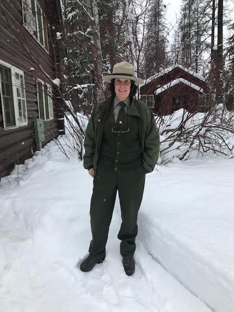 Laura Law Woman ranger in uniform stands in snow next to log cabin building
