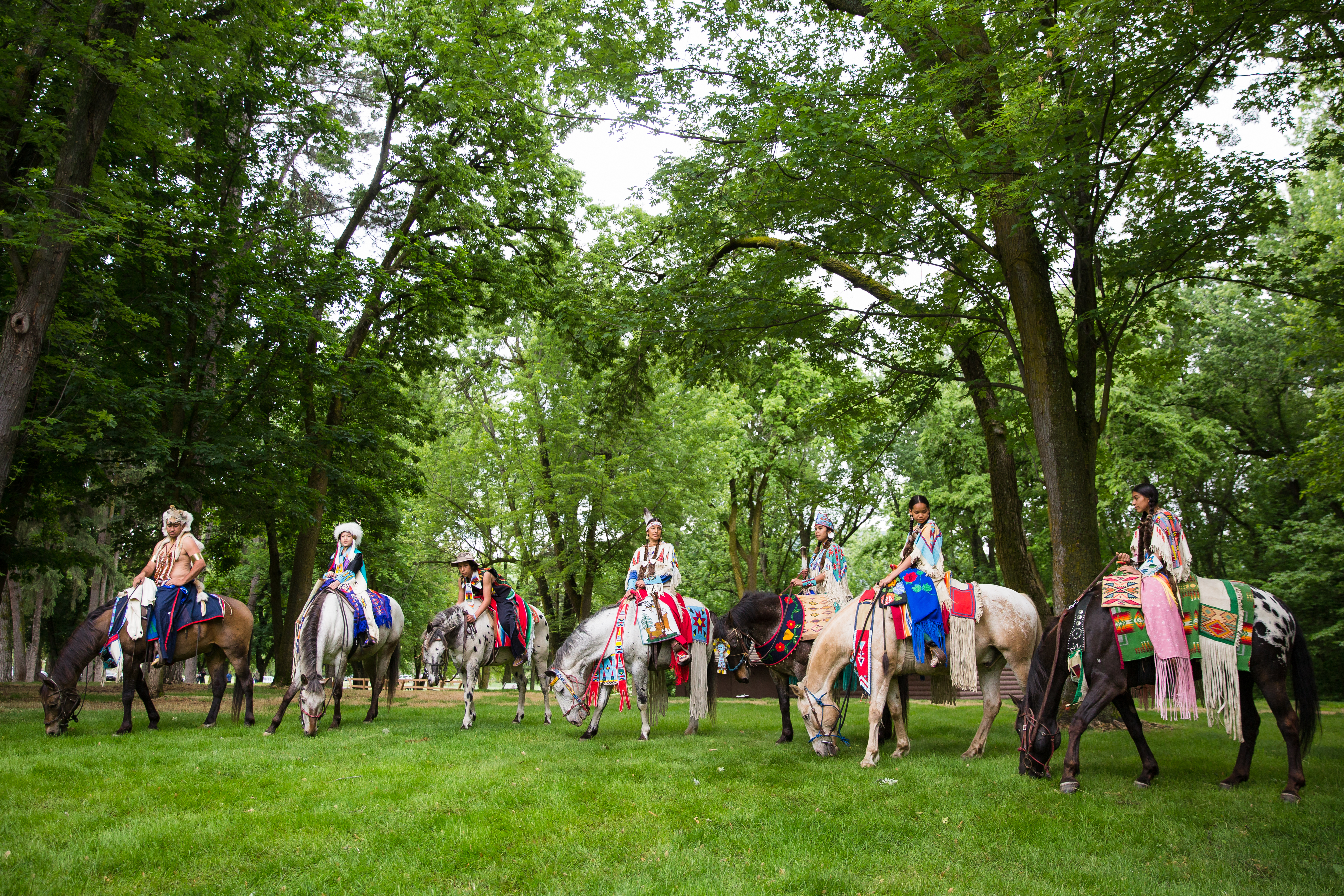 Seven horses and riders in traditional dress with green grass and trees surrounding them.