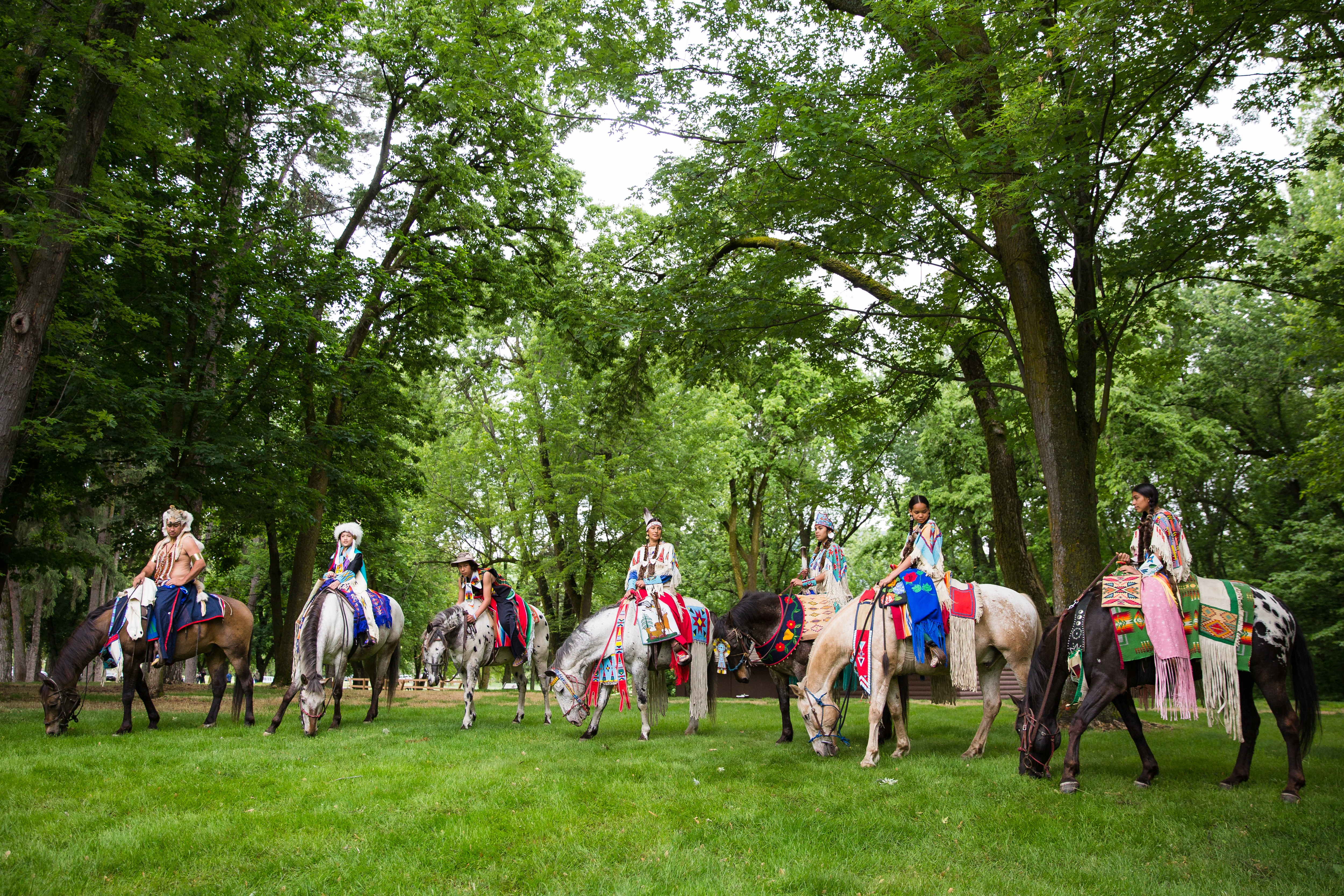 Nez Perce Horse Parade Seven horses and riders in traditional dress with green grass and trees surrounding them.