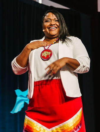 Tai Simpson Native woman standing on a stage wearing a white shirt, a red skirt with ribbons of various colors on the bottom, and a large beaded medallion around her neck.