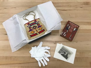 Collections talk A colorful cornhusk bag sits on white tissue paper near a pair of white gloves, a book, and black and white photographs.