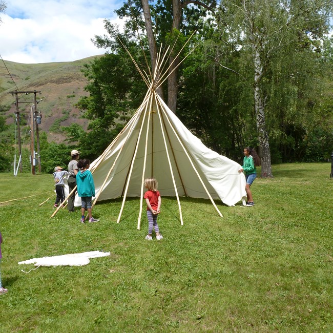 A ranger shows four children how to put a tipi up in a grassy park using bare tipi poles and a white canvas