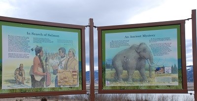 A sign with two informational panels about Tolo Lake is located on the shoreline of a lake.