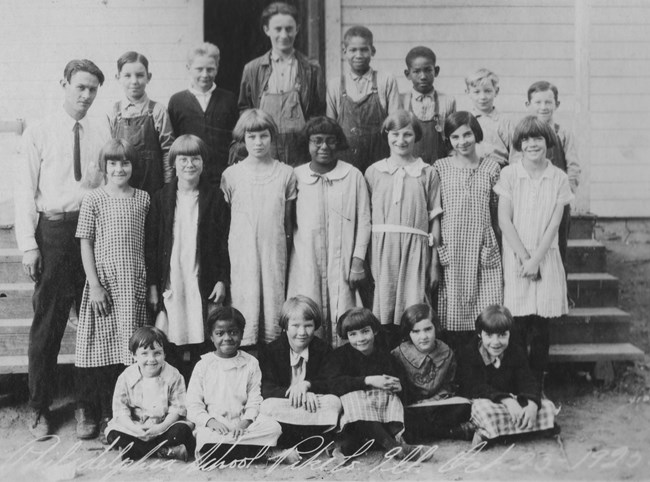 Historical photo of 20 children of mixed ages and genders posing for the camera in three rows in front of the steps to a building. The group appears to be 20% Black and 80% White. A female teacher stands in the middle of the back row.